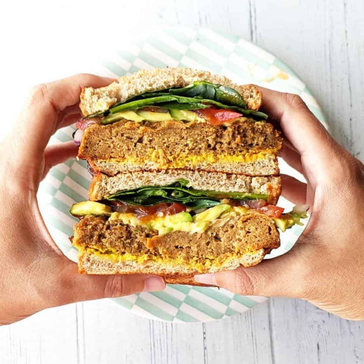 hands holding a sandwich filled with seitan and veggies.